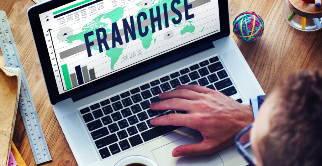 A Man with a Mug of Coffee in His Left Hand and His Right Hand on a Laptop Keyboard with a Franchise Graphic on the Screen