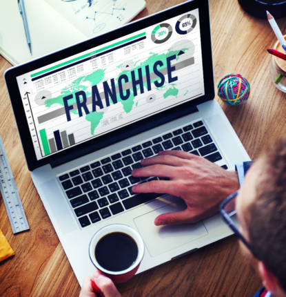 A Man with a Mug of Coffee in His Left Hand and His Right Hand on a Laptop Keyboard with a Franchise Graphic on the Screen