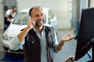 A smiling man on a cell phone stands in an auto repair bay with a mechanic and car behind him and looks at a computer screen